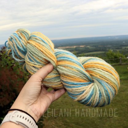 A hand holds a skein of thick, multicolored yarn in shades of yellow, blue, and light green. The background features a lush, green landscape and an expansive, cloudy sky. The text "LEILANI HANDMADE" is faintly visible at the bottom of the image.