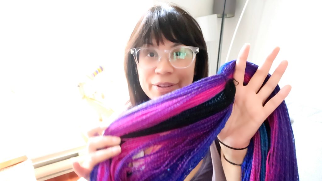 A woman wearing glasses holds out a colorful yarn with the colors purple, pink, black and blue. She spreads the skein out to show the detail. The yarn was spun using the Lendrum fast flyer.