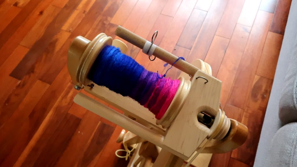 A bobbin is half full of handspun singles on a spinning wheel. The photo is shot from above with a close up of the Lendrum fast flyer. The colors of the yarn are blue purple and pink.