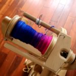 A bobbin is half full of handspun singles on a spinning wheel. The photo is shot from above with a close up of the Lendrum fast flyer. The colors of the yarn are blue purple and pink.