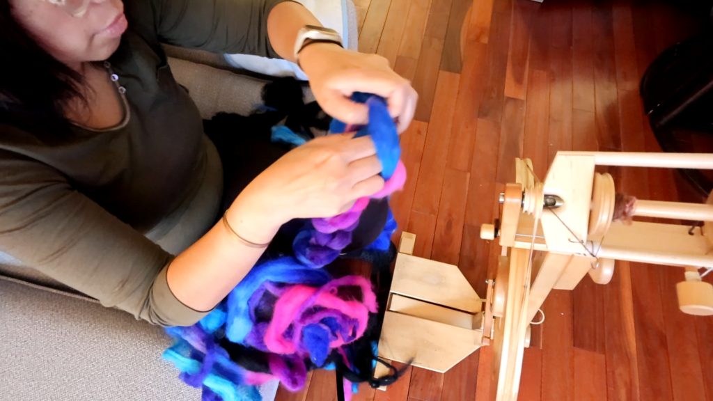 A person sitting on a couch holding a blue and pink roving in front of a spinning wheel, getting ready to try out the Lendrum fast flyer.
