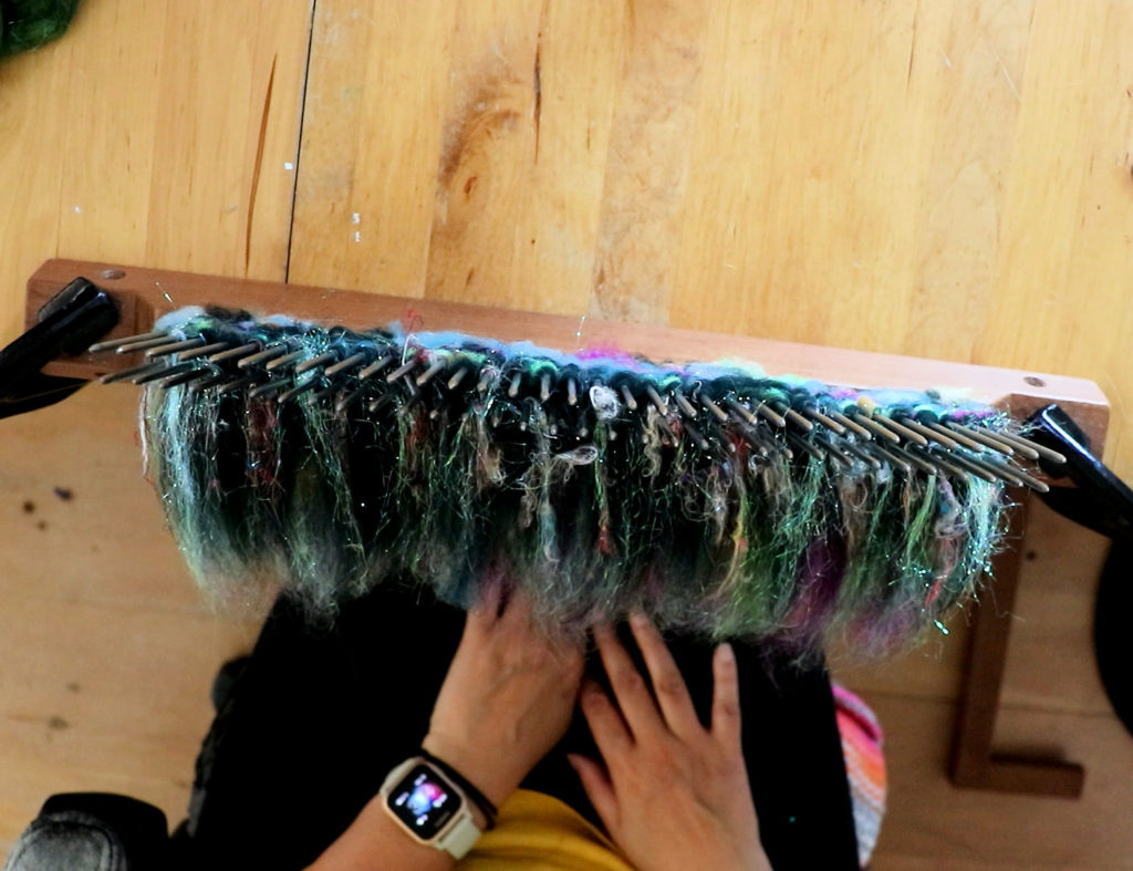 A person wearing a smartwatch sits at a wooden table, using a blending hackle to blend colorful fibers. Their hands are visible near the fibers, which are a mix of green, purple, blue, and shiny strands, all attached to metal spikes of the tool.