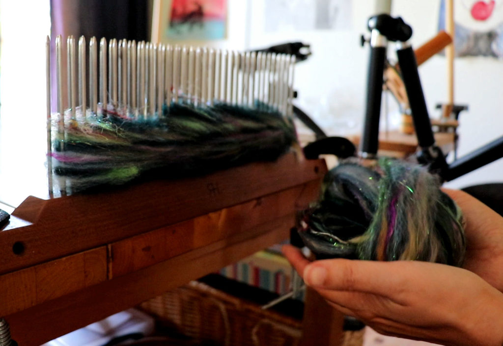 A person holds multicolored, shiny fiber near a wooden blending board with metal pins. More fiber is spread across the board’s pins. The background shows a blurred room with art, books, and weaving tools. Light softly enters from the left.