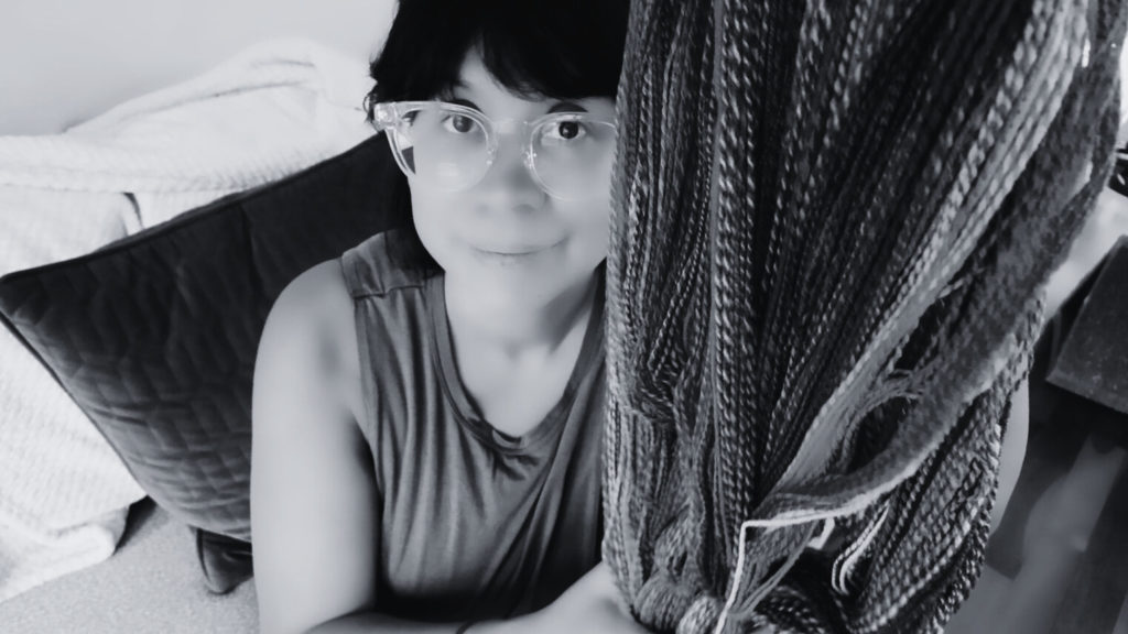 A person with short dark hair and clear glasses, wearing a sleeveless shirt, looks up at the camera while holding a large untwisted skein of handspun yarn. The image is in black and white, with a couch and blanket in the background. The image is in black and white so not to give away the final process from spinning the roving.