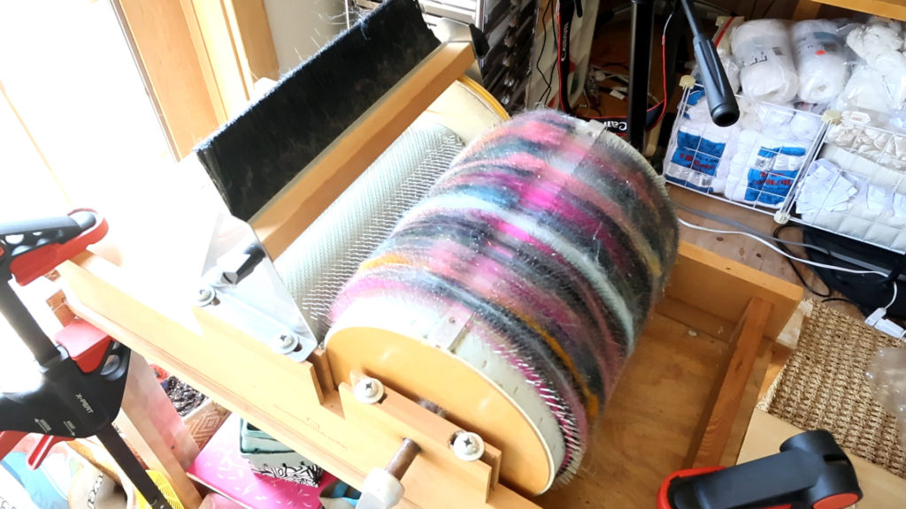 A drum carder loaded with colorful, blended fibers in shades of pink, purple, white, orange, and black sits on a wooden table. Various crafting supplies and equipment are visible in the background.