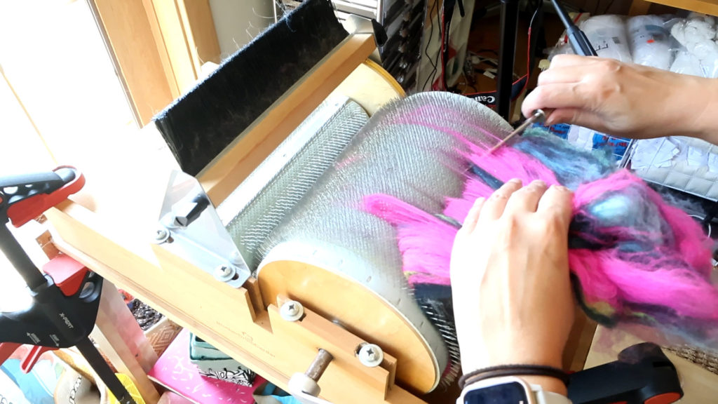 A person uses a drum carder to blend pink, blue, and black fibers for spinning. Their hands guide the fibers off the carding drum using a doffer to remove the fibers. Sunlight streams in from the left, and various tools and yarn are visible in the background. The person wears a smartwatch.
