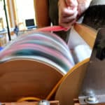 A close-up of a person using a fiber carding machine with a spinning drum. The person holds pink fiber against the drum, blending colorful wool fibers. Workshop tools, and daylight are visible in the blurred background.