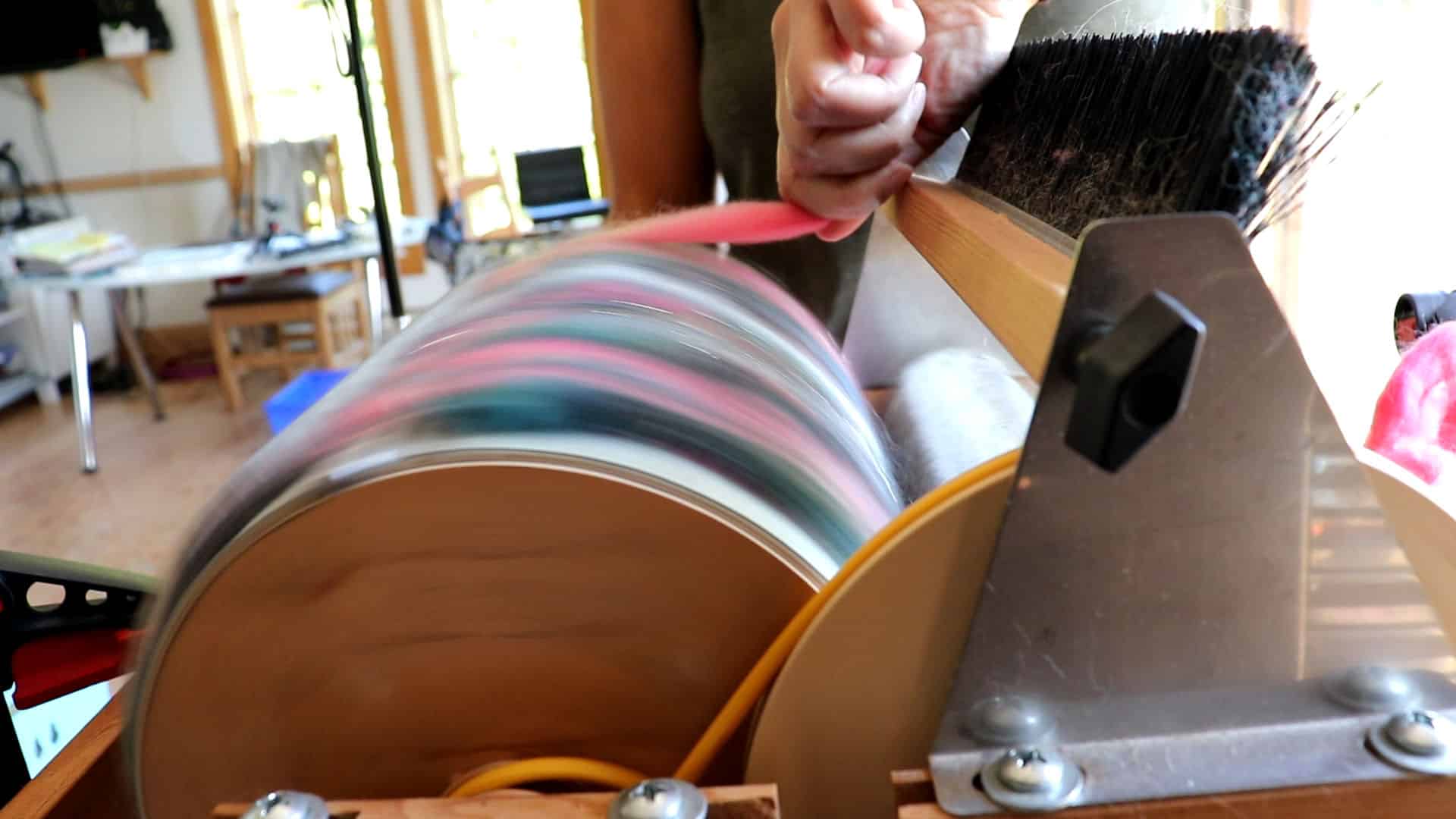 A close-up of a person using a fiber carding machine with a spinning drum. The person holds pink fiber against the drum, blending colorful wool fibers. Workshop tools, and daylight are visible in the blurred background.
