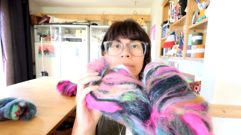 A person with glasses and short dark hair holds up a colorful wool batt with pink, blue, green, and black fibers in a bright, cozy craft room filled with shelves, supplies, and a wooden table.