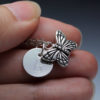 A close-up of a hand holding a silver necklace with a round charm engraved with the letter "B" and a detailed butterfly pendant. The background is softly blurred, and "LEILANI HANDMADE" is faintly visible at the bottom.