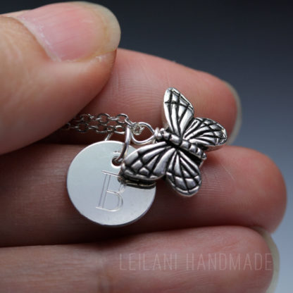 A close-up of a hand holding a silver necklace with a round charm engraved with the letter "B" and a detailed butterfly pendant. The background is softly blurred, and "LEILANI HANDMADE" is faintly visible at the bottom.