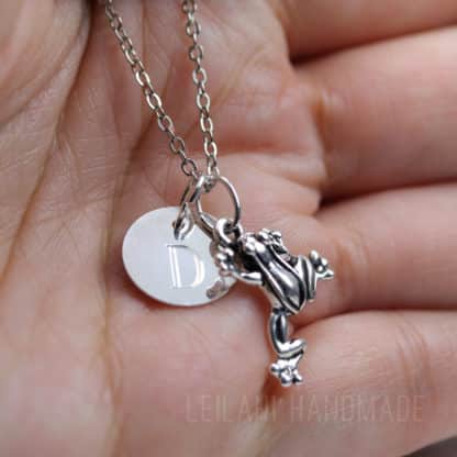 A close-up of a silver necklace with two charms resting on an open palm: a round disc engraved with the letter "D" and a small, detailed frog figurine. The image has a soft, natural lighting and the text “LEILANI HANDMADE” is subtly visible at the bottom.