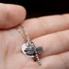 A close-up of a hand holding a delicate silver necklace with a dragonfly charm and a round disc pendant. The background is blurred, and the chain is draped gently across the palm. The text "LEILANI HANDMADE" is faintly visible at the bottom right.