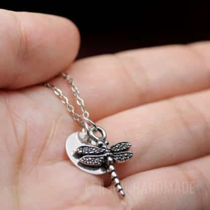 A close-up of a hand holding a delicate silver necklace with a dragonfly charm and a round disc pendant. The background is blurred, and the chain is draped gently across the palm. The text "LEILANI HANDMADE" is faintly visible at the bottom right.