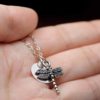 A close-up of a hand holding a delicate silver necklace with a dragonfly charm and a round disc pendant. The background is blurred, and the chain is draped gently across the palm. The text "LEILANI HANDMADE" is faintly visible at the bottom right.