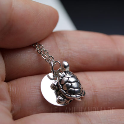 A close-up of a hand holding a silver necklace with a small, detailed turtle charm and a round flat pendant. The turtle has a textured shell and tiny legs. The background is blurred, drawing attention to the jewelry.