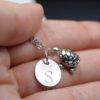 A hand holds a silver necklace featuring a round charm stamped with the letter “S” and a small, detailed turtle charm. The background is softly blurred, and the text “LEILANI HANDMADE” is faintly visible at the bottom.