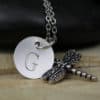 A silver necklace features a round pendant engraved with the letter "G" and a detailed dragonfly charm. Both pendants hang from a silver chain, with a smooth black stone and a blurred "Leilani Handmade" watermark in the background.