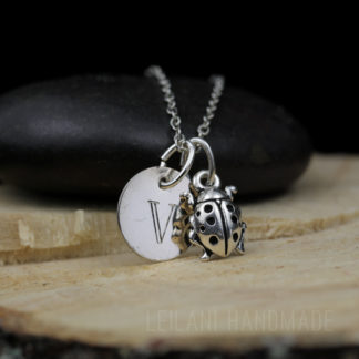 A silver ladybug necklace features a round pendant engraved with the letter "V" and a detailed ladybug charm. Both pendants hang from a delicate chain, draped over a smooth black stone on a wooden surface. The text "LEILANI HANDMADE" is faintly visible.