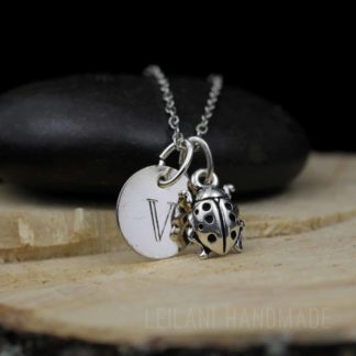 A silver ladybug necklace features a round pendant engraved with the letter "V" and a detailed ladybug charm. Both pendants hang from a delicate chain, draped over a smooth black stone on a wooden surface. The text "LEILANI HANDMADE" is faintly visible.