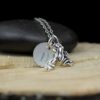 A silver necklace with a frog charm and a round pendant engraved with the letter "D" rests on a smooth black stone atop a light wooden surface. The background is dark, highlighting the details of the jewelry.