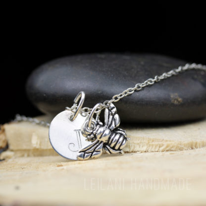 A silver necklace with a round pendant engraved with the letter "J" and a detailed bee charm rests on a smooth black stone, which is placed on a light wooden surface. The background is softly blurred with "LEILANI HANDMADE" faintly visible.