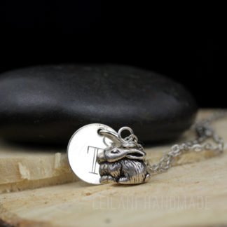 A silver necklace features a round disc engraved with the letter "T" and a detailed bunny rabbit charm. The necklace rests on cut wood, with a smooth black stone in the background. The text "LEILANI HANDMADE" is faintly visible at the bottom.
