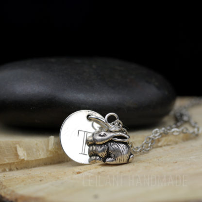 A silver necklace features a round disc engraved with the letter "T" and a detailed bunny rabbit charm. The necklace rests on cut wood, with a smooth black stone in the background. The text "LEILANI HANDMADE" is faintly visible at the bottom.