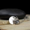 A silver necklace features a round disc engraved with the letter "T" and a detailed bunny rabbit charm. The necklace rests on cut wood, with a smooth black stone in the background. The text "LEILANI HANDMADE" is faintly visible at the bottom.