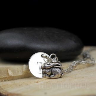 A silver necklace features a round disc engraved with the letter "T" and a detailed bunny rabbit charm. The necklace rests on cut wood, with a smooth black stone in the background. The text "LEILANI HANDMADE" is faintly visible at the bottom.