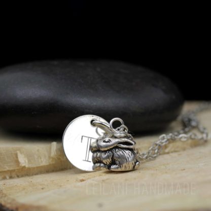 A silver necklace features a round disc engraved with the letter "T" and a detailed bunny rabbit charm. The necklace rests on cut wood, with a smooth black stone in the background. The text "LEILANI HANDMADE" is faintly visible at the bottom.