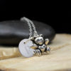 A silver necklace features a round pendant engraved with the letter "R" and a small, detailed teddy bear charm. Both pendants rest on a wooden surface with a smooth, dark stone in the background. The text "LEILANI HANDMADE" is faintly visible below.