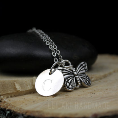 A silver necklace with a butterfly charm and a round pendant engraved with the letter "C," displayed on a smooth black stone and a wooden slab. The engraving "LEILANI HANDMADE" is faintly visible at the bottom of the image.