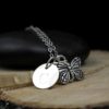 A silver necklace with a butterfly charm and a round pendant engraved with the letter "C," displayed on a smooth black stone and a wooden slab. The engraving "LEILANI HANDMADE" is faintly visible at the bottom of the image.
