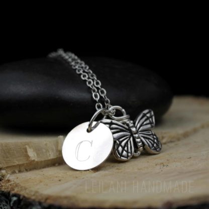 A silver necklace with a butterfly charm and a round pendant engraved with the letter "C," displayed on a smooth black stone and a wooden slab. The engraving "LEILANI HANDMADE" is faintly visible at the bottom of the image.