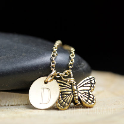 A gold necklace with a round pendant engraved with the letter "D" and a detailed butterfly charm rests on a smooth black stone, with a light-colored textured surface beneath and a dark background.