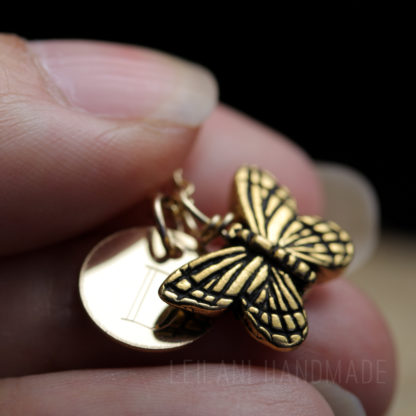 A close-up of fingers holding a gold butterfly charm next to a round gold tag engraved with the letter D. The butterfly has detailed wings, and both charms have a polished, metallic finish. The background is softly blurred.