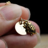 A close-up of fingers holding a gold necklace with two charms: a round disc engraved with a letter E, and a detailed ladybug pendant. The background is dark and out of focus, and the text "LEILANI HANDMADE" appears faintly at the bottom.