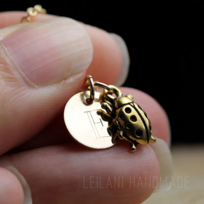 A close-up of fingers holding a gold necklace with two charms: a round disc engraved with a letter E, and a detailed ladybug pendant. The background is dark and out of focus, and the text "LEILANI HANDMADE" appears faintly at the bottom.