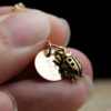 A close-up of fingers holding a gold necklace with two charms: a round disc engraved with a letter E, and a detailed ladybug pendant. The background is dark and out of focus, and the text "LEILANI HANDMADE" appears faintly at the bottom.