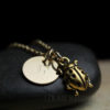 A gold necklace with a circular pendant engraved with the letter "E" and a small, detailed ladybug charm rests on a smooth black stone. The background is dark, highlighting the shiny metal and the engraved text "LEILANI HANDMADE.