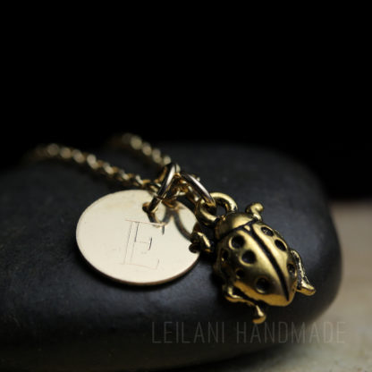 A gold necklace with a circular pendant engraved with the letter "E" and a small, detailed ladybug charm rests on a smooth black stone. The background is dark, highlighting the shiny metal and the engraved text "LEILANI HANDMADE.