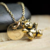 A close-up of a gold necklace featuring a round charm engraved with the letter "S" and a small gold teddy bear pendant. The jewelry is set against a smooth stone and wooden surface. The words "LEILANI HANDMADE" are faintly visible at the bottom.