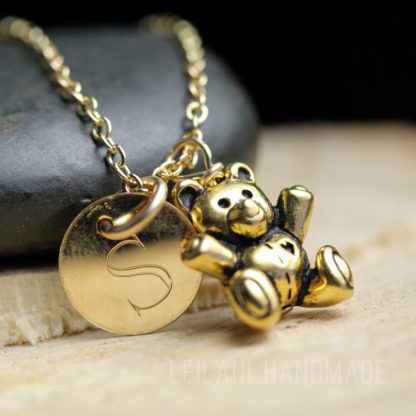 A close-up of a gold necklace featuring a round charm engraved with the letter "S" and a small gold teddy bear pendant. The jewelry is set against a smooth stone and wooden surface. The words "LEILANI HANDMADE" are faintly visible at the bottom.