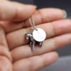 A hand holds a silver necklace with two charms: one is a small, detailed horse and the other is a round disc engraved with the letter “E.” The background is blurred and the watermark “LEILANI HANDMADE” is visible at the bottom.