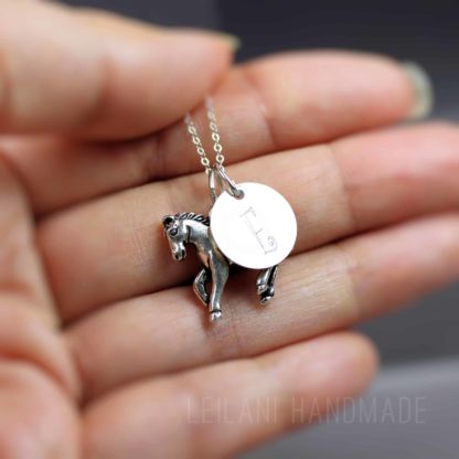 A hand holds a silver necklace with two charms: one is a small, detailed horse and the other is a round disc engraved with the letter “E.” The background is blurred and the watermark “LEILANI HANDMADE” is visible at the bottom.