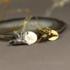 Two hummingbird keepsake necklace, with hummingbird charms, one silver with an "M" engraved charm and one gold with an "H" engraved charm, rest on a smooth stone. Both are on delicate chains. The background is blurred, and "LEILANI HANDMADE" is faintly visible at the bottom.