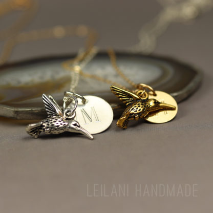 Two hummingbird keepsake necklace, with hummingbird charms, one silver with an "M" engraved charm and one gold with an "H" engraved charm, rest on a smooth stone. Both are on delicate chains. The background is blurred, and "LEILANI HANDMADE" is faintly visible at the bottom.