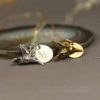 Two hummingbird keepsake necklace, with hummingbird charms, one silver with an "M" engraved charm and one gold with an "H" engraved charm, rest on a smooth stone. Both are on delicate chains. The background is blurred, and "LEILANI HANDMADE" is faintly visible at the bottom.