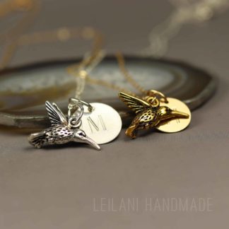 Two hummingbird keepsake necklace, with hummingbird charms, one silver with an "M" engraved charm and one gold with an "H" engraved charm, rest on a smooth stone. Both are on delicate chains. The background is blurred, and "LEILANI HANDMADE" is faintly visible at the bottom.